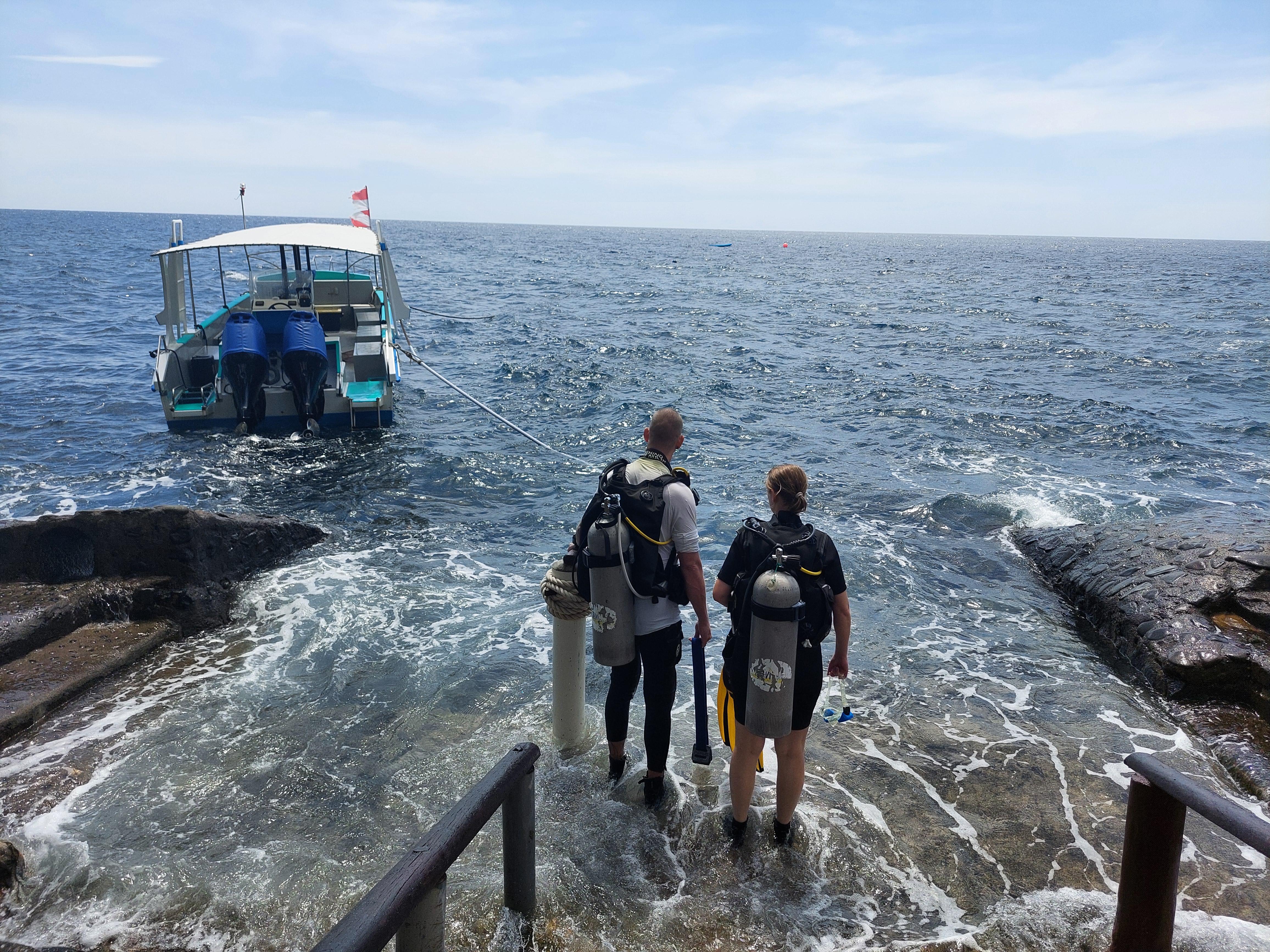Zwei Taucher betreten das Meer von einer Felsplattform aus, ein Boot wartet im klaren blauen Wasser.