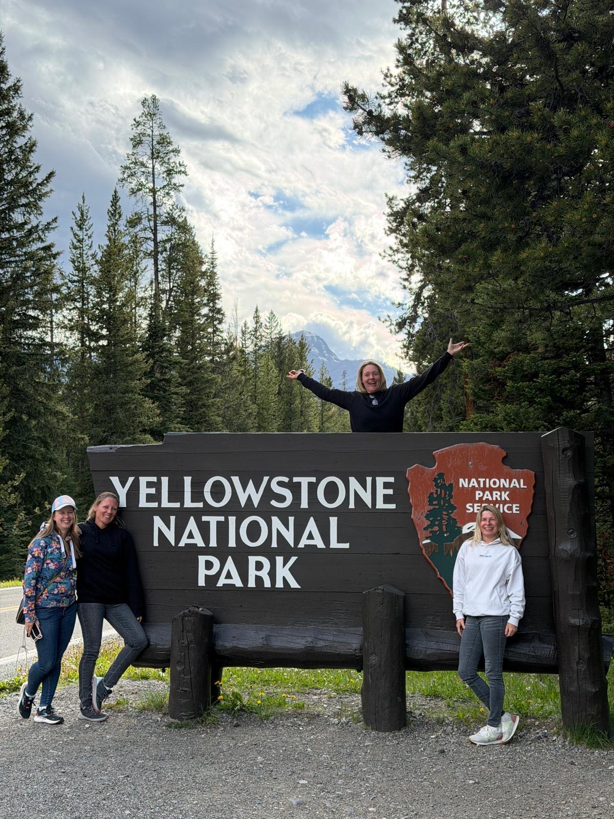 Vier Personen vor dem großen Schild des Yellowstone Nationalparks, umgeben von hohen Kiefern.