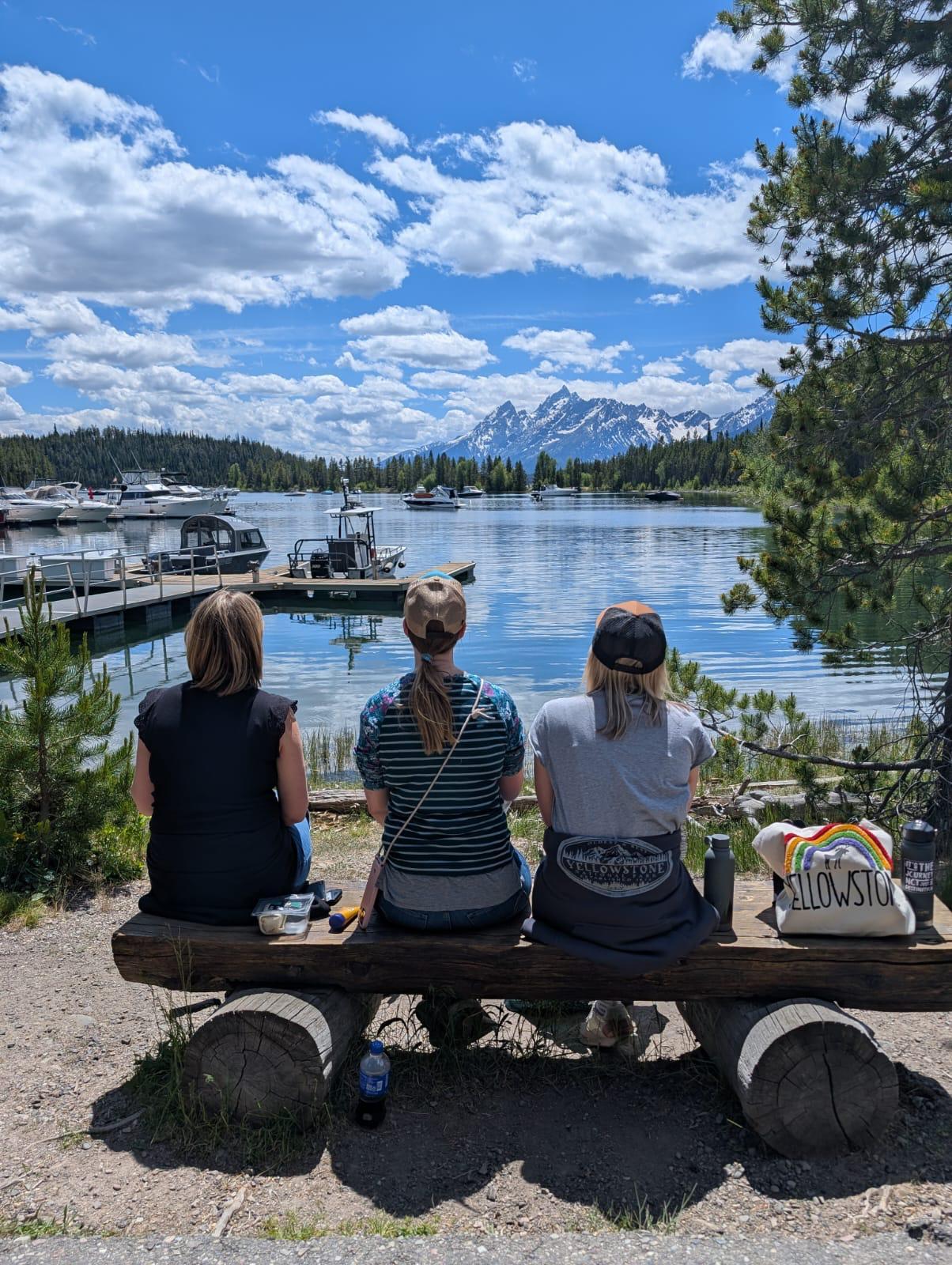 Drei Personen sitzen auf einer Holzbank am See mit Blick auf Boote und bergige Landschaft unter blauem Himmel.
