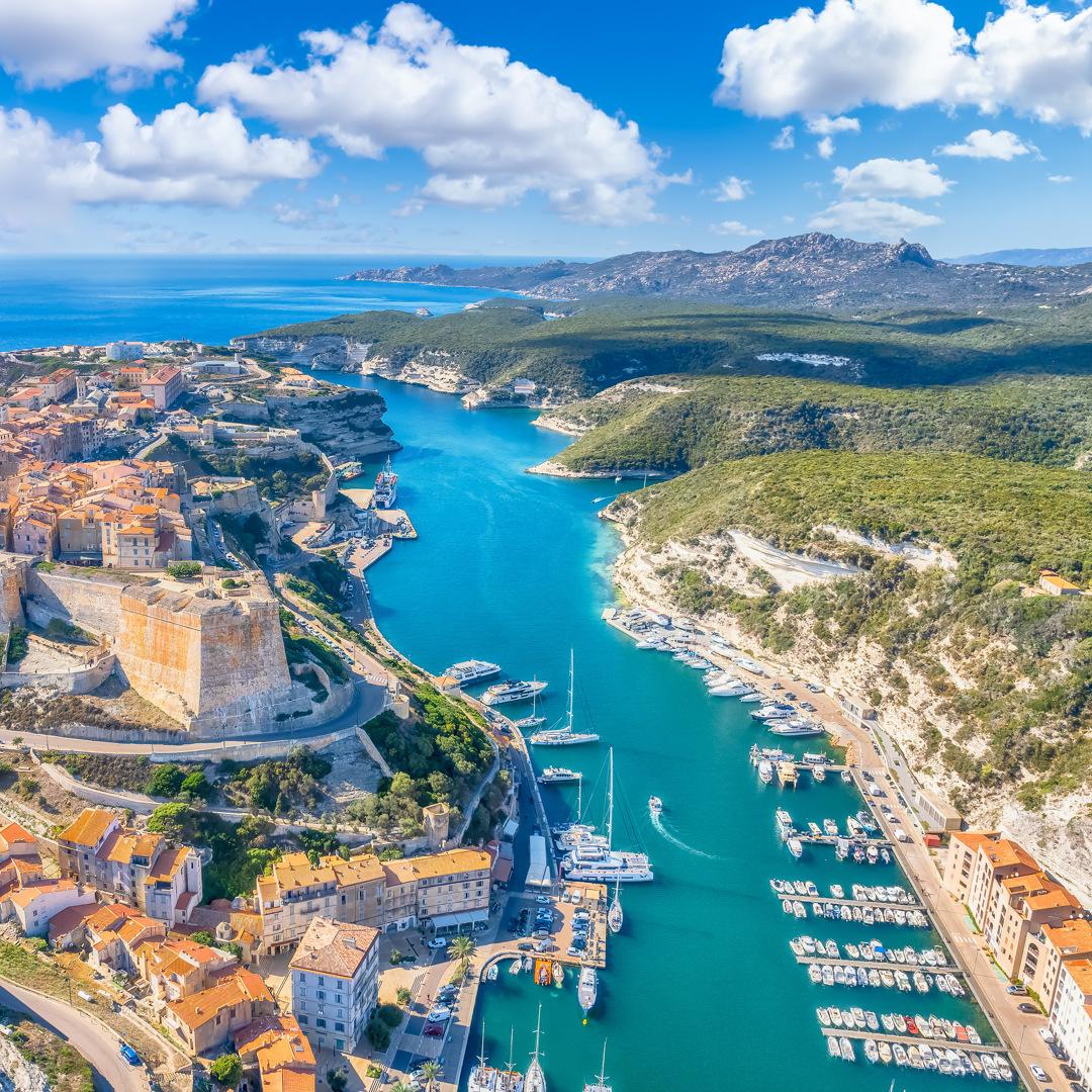 Bucht mit klarem Wasser, Yachten und historischer Stadt umgeben von grünen Hügeln unter blauem Himmel.
