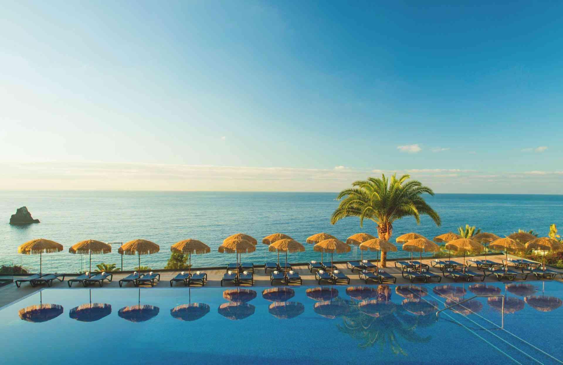 Luxushotel mit großem Pool, Liegestühlen, Sonnenschirmen und Meerblick unter klarem Himmel.