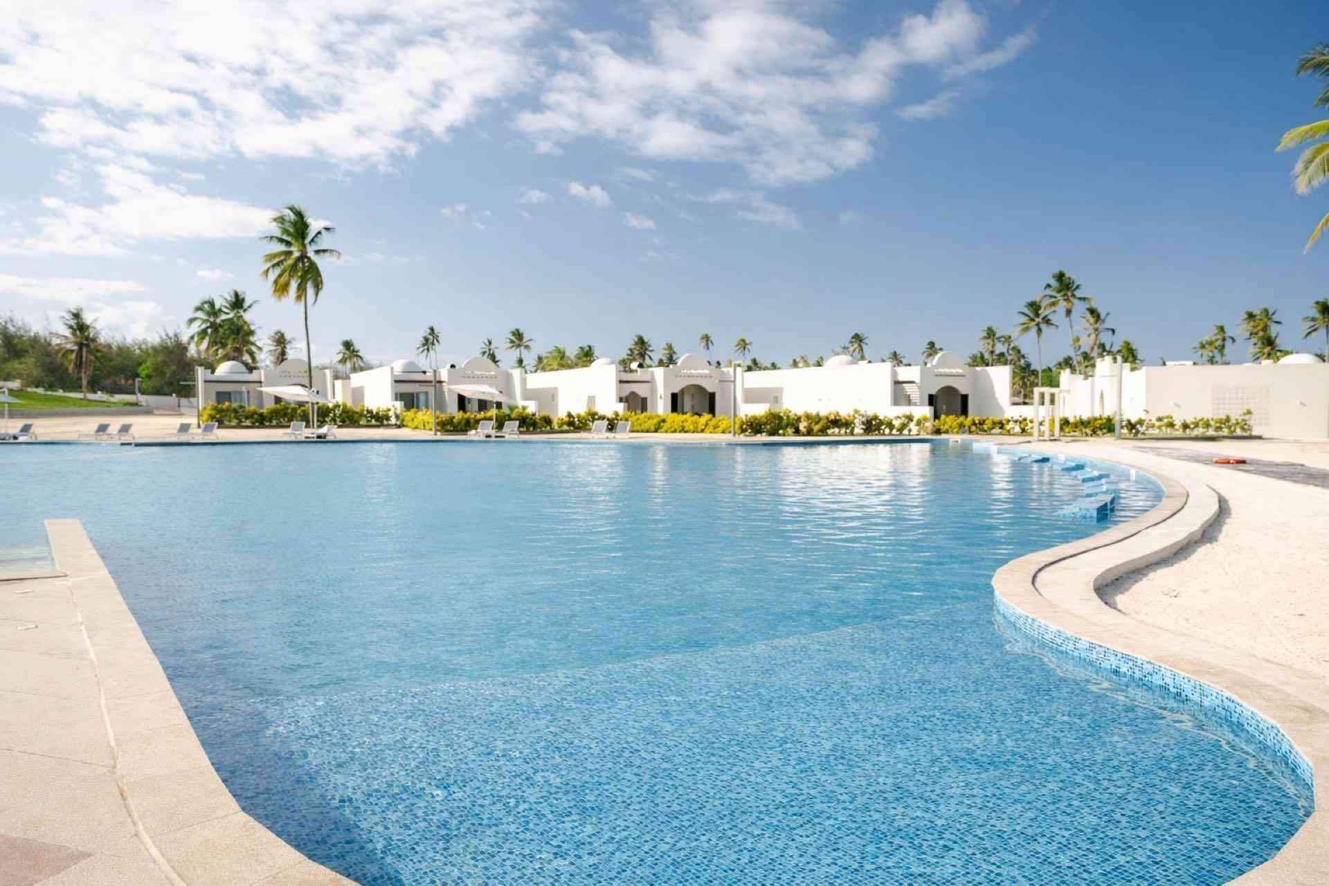 Großer, geschwungener Hotelpool mit kristallklarem Wasser, weißem Gebäude und Palmen unter blauem Himmel.