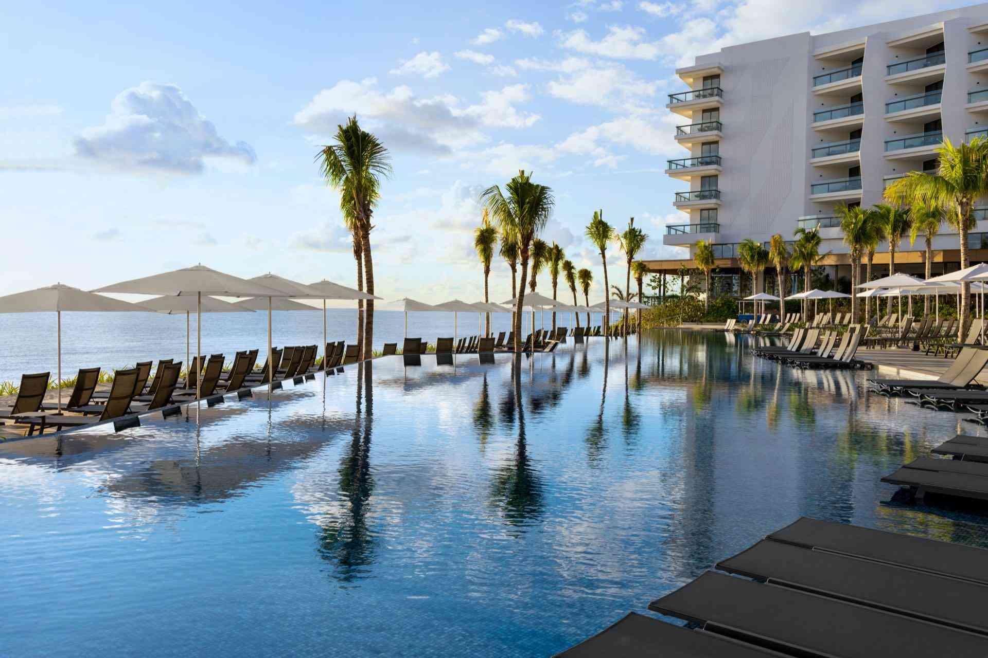 Ein moderner Hotelpool mit Liegestühlen, Sonnenschirmen und Palmen, Blick auf Meer und Berge.
