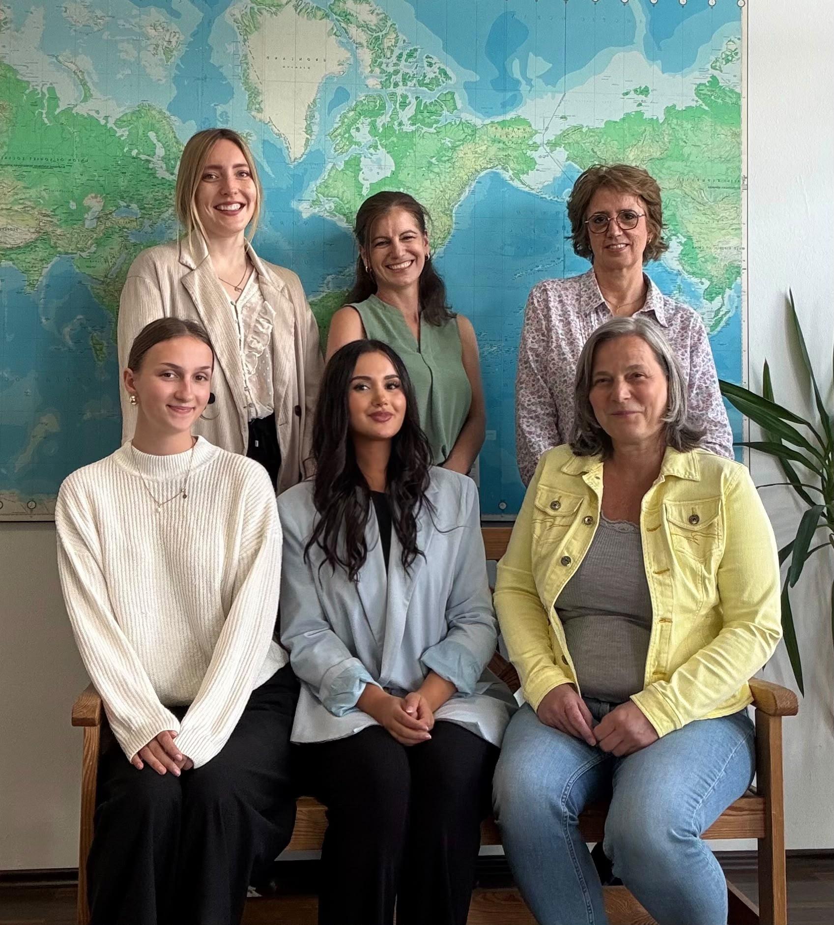 Sechs Frauen vor großer Weltkarte, entspannte Kleidung, freundlich, Reisebüro oder Hotelteam.