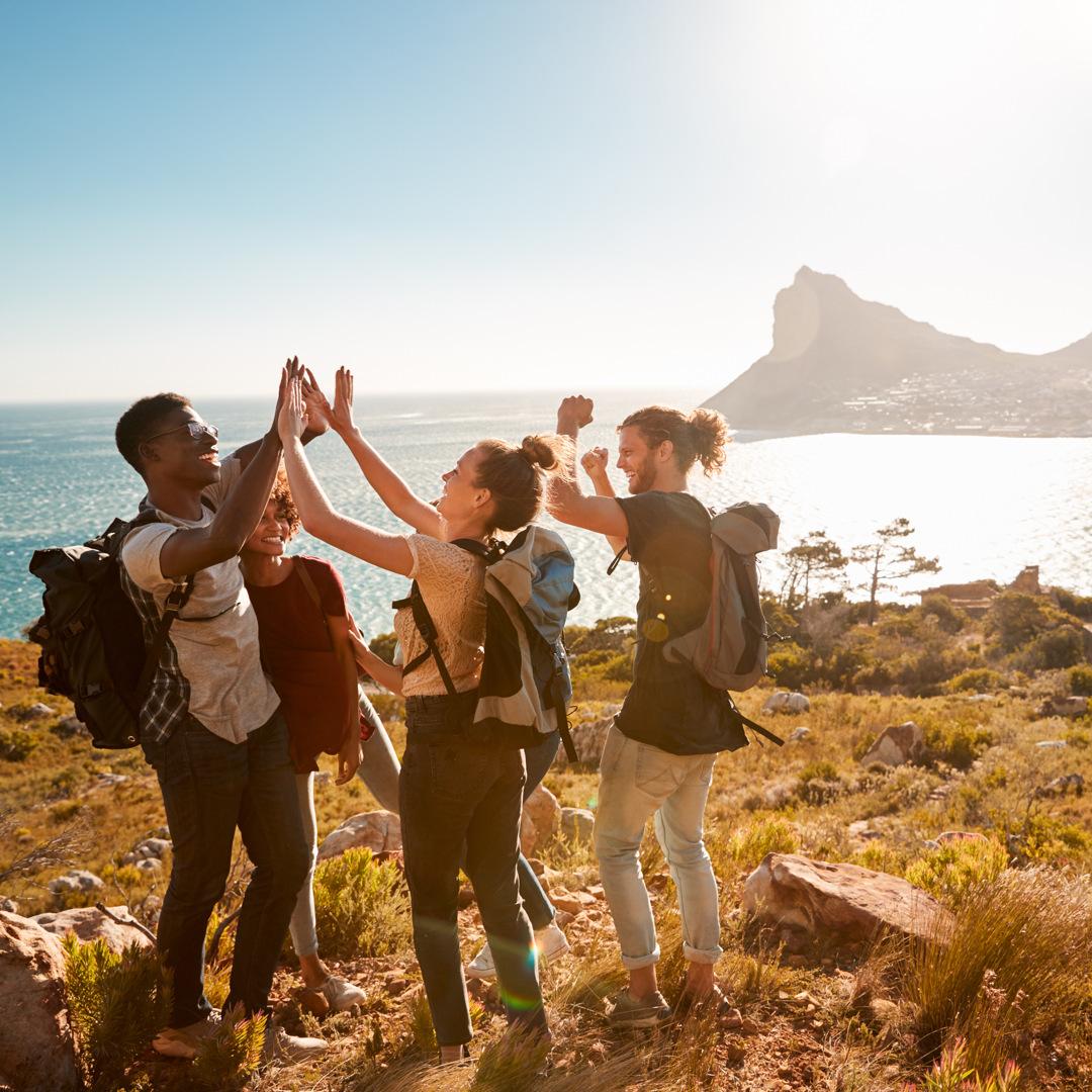 Eine Gruppe junger Reisender genießt auf einer sonnigen Klippe mit Berg- und Meerblick den Urlaub.