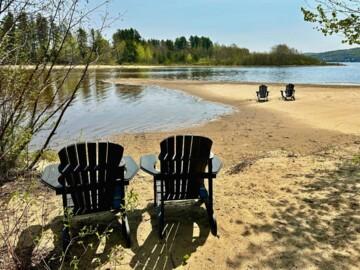 Zwei Holzstühle am ruhigen Sandstrand eines Sees, umgeben von Bäumen und klarer blauer Himmel.