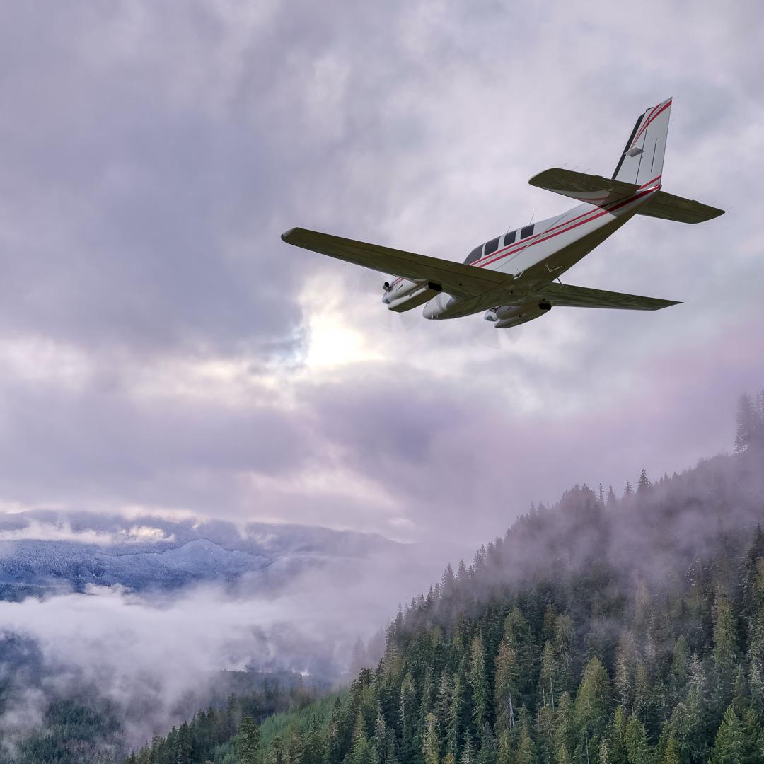 Kleines Flugzeug fliegt über nebelverhangene, bewaldete Berge bei bewölktem Himmel.