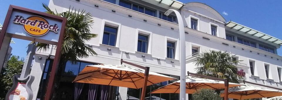 Außenansicht eines weißen Gebäude mit Hard Rock Café und orangenen Sonnenschirmen, Palmen davor.