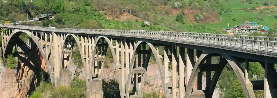 Massive Betonbogenbrücke über eine tiefe Schlucht, umgeben von grüner Landschaft und kleinen Häusern.
