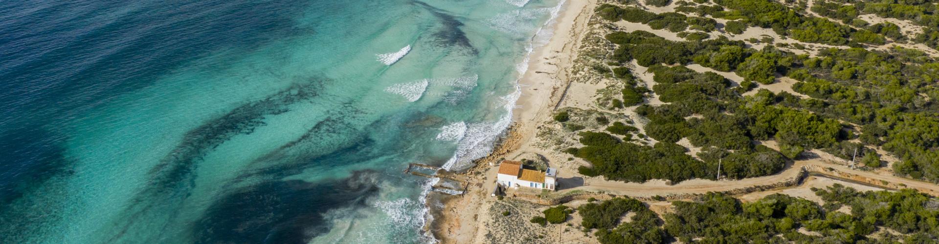 Türkisblaues Meer trifft auf sandigen Küstenabschnitt mit grün bewachsenem Hügel und kleinem Gebäude.