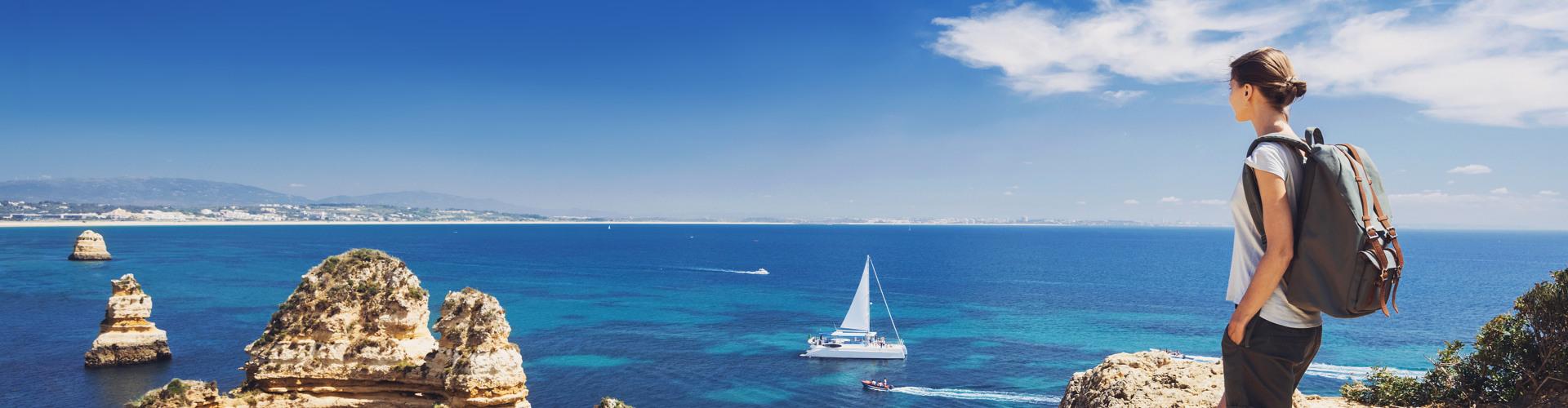 Frau mit Rucksack blickt von Klippe auf Segelboot und tiefblaues Meer unter blauem Himmel.