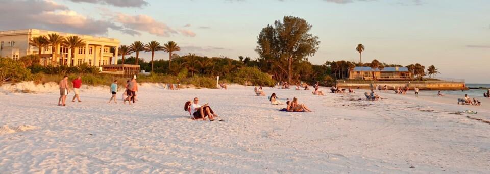Weißer Sandstrand mit Hotelgebäuden, Palmen und entspannten Urlaubern bei Sonnenuntergang.