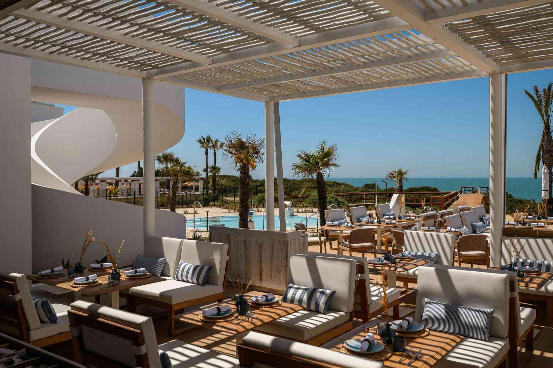 Offene Poolbar mit modernen Holzmöbeln, Palmendekor und blick auf das blaue Meer.