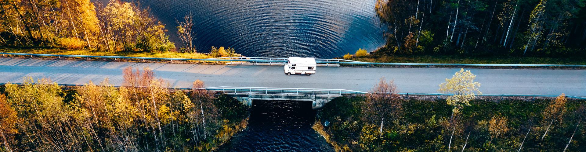 Wohnmobil fährt auf Brücke über ruhigen See, umgeben von herbstlich buntem Wald, ideal für Urlaub.