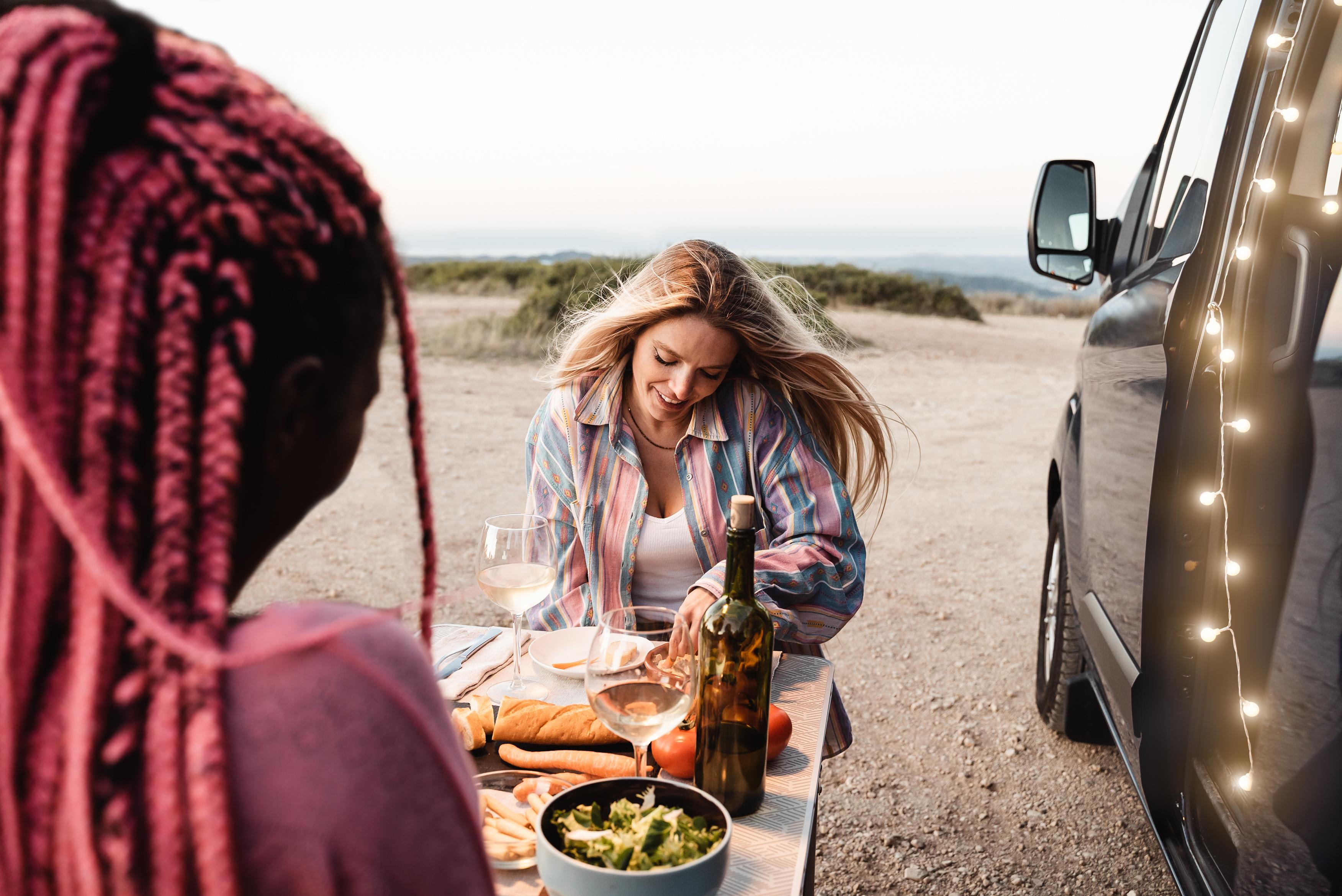 Zwei Frauen genießen ein Picknick am Camper mit Meerblick bei Sonnenuntergang.