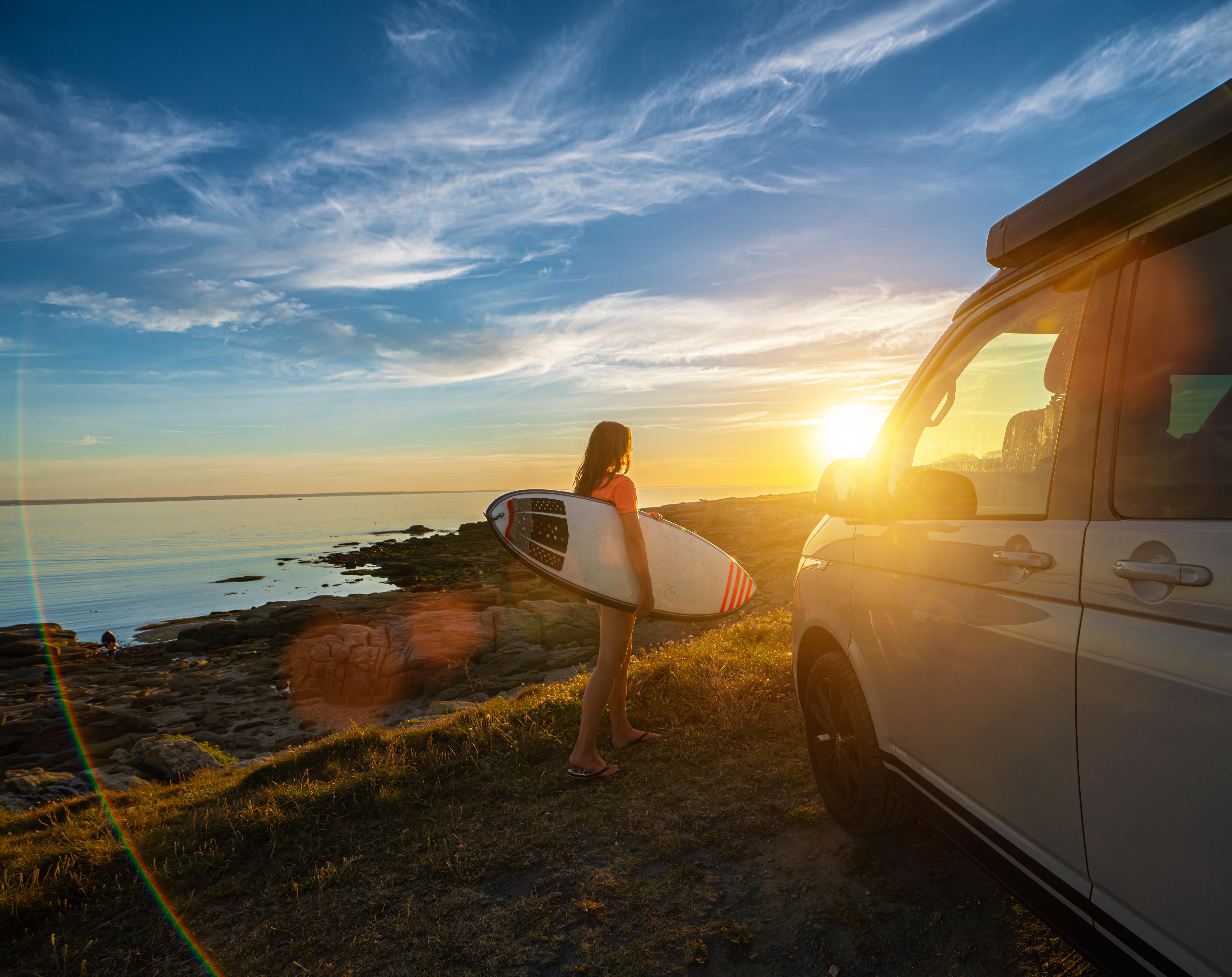 Frau mit Surfbrett am Meeresufer bei Sonnenuntergang, neben einem weißen Campervan auf dem Gras.