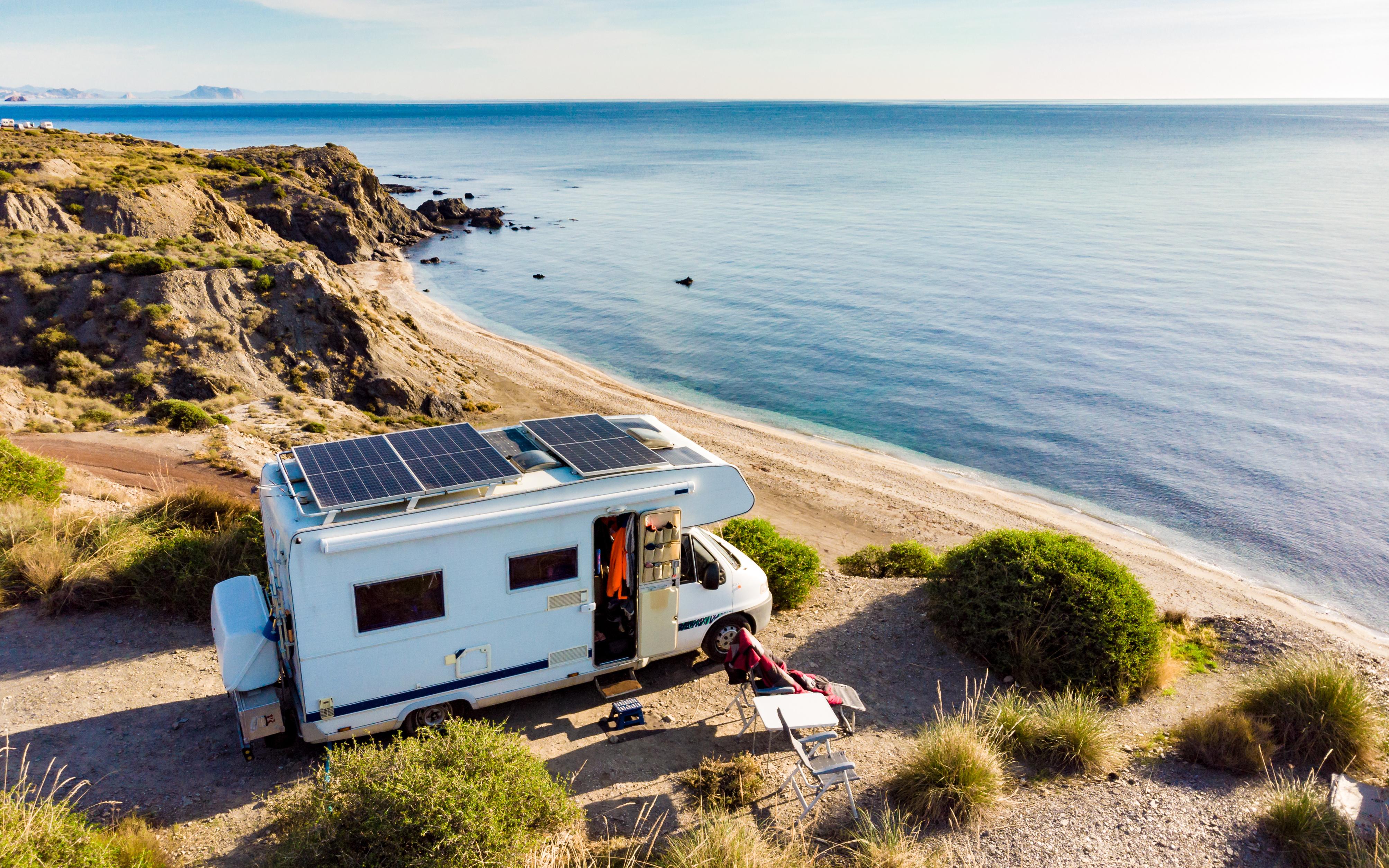 Wohnmobil mit Solarpanelen steht ruhig am Küstenstrand mit blauem Meer und felsiger Klippe.