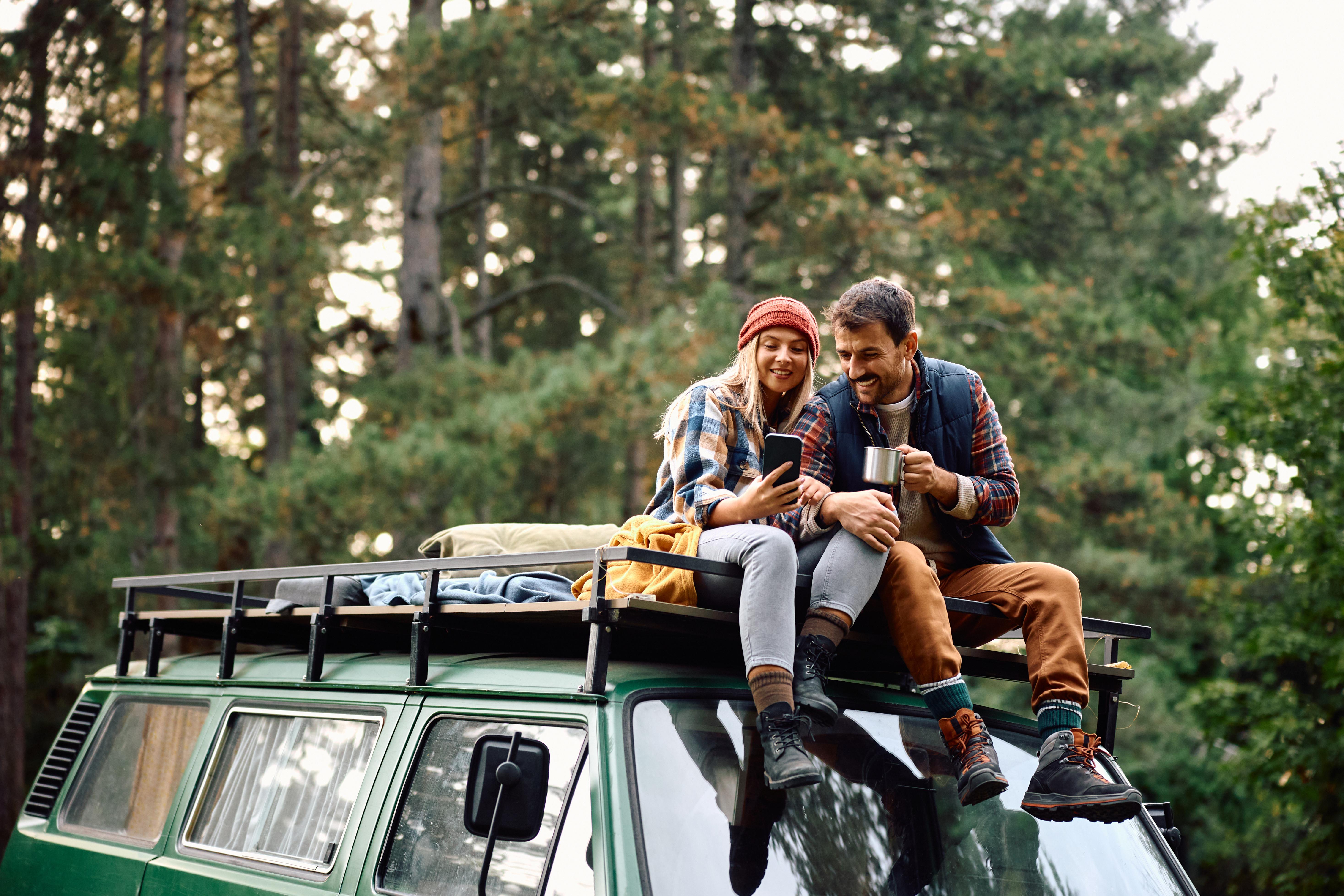 Pärchen sitzt auf Van-Dach im Wald, trinkt Kaffee, entspannt auf Outdoor-Reise oder Campingurlaub.